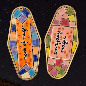 May include: Two decorative keychains with a mosaic design. One keychain has the text "made of magic" on an orange background. The other keychain has the text "everyday magic" on a pink background. Both keychains have a gold-colored border.