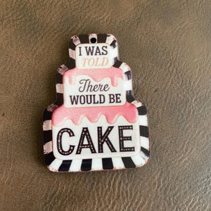 May include: A cake-shaped decorative item with the text "I WAS TOLD There WOULD BE CAKE." The item has a pink frosting layer and a black and white striped border. The background is a textured brown surface.