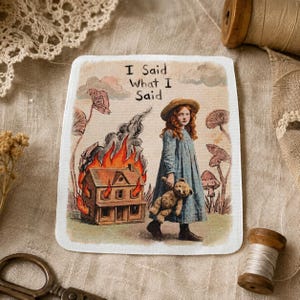 May include: A rectangular fabric patch featuring a printed illustration of a red-haired child in a blue dress holding a teddy bear. Beside the child, a house is engulfed in orange flames. The text "I Said What I Said" is printed above the scene.