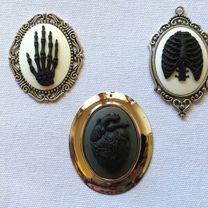 May include: Three silver and gold framed cameo pendants with black anatomical images. The pendants feature a skeleton hand, a ribcage, and a heart.