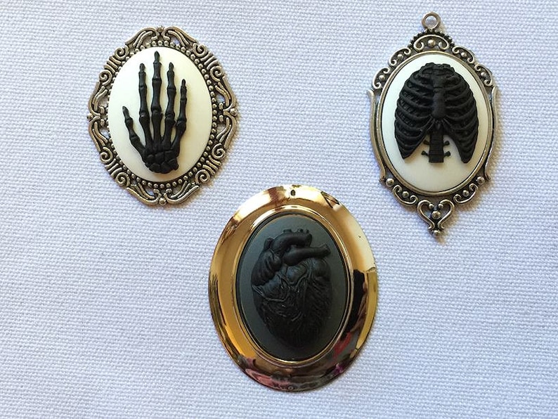 May include: Three silver and gold framed cameo pendants with black anatomical images. The pendants feature a skeleton hand, a ribcage, and a heart.