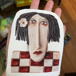 May include: A white ceramic sculpture of a woman's face with a red and white checkered background. The woman has long black hair, a white flower behind her ear, and a long nose.