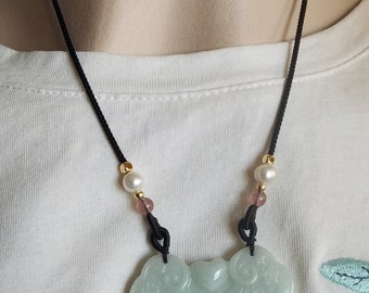 Puede incluir: Collar con colgante de jade y cordón negro. El colgante verde claro presenta un diseño floral tallado. El collar también incluye perlas, cuentas rosas y detalles dorados.