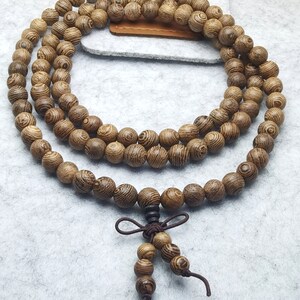 Puede incluir: Un collar de cuentas de oraci&oacute;n de madera con un acabado marr&oacute;n oscuro. Las cuentas son redondas y tienen un patr&oacute;n de vetas de madera visible. Una borla de cuentas m&aacute;s peque&ntilde;as y un cord&oacute;n anudado est&aacute;n unidos a la parte inferior del collar. Las cuentas se muestran sobre una superficie gris claro.
