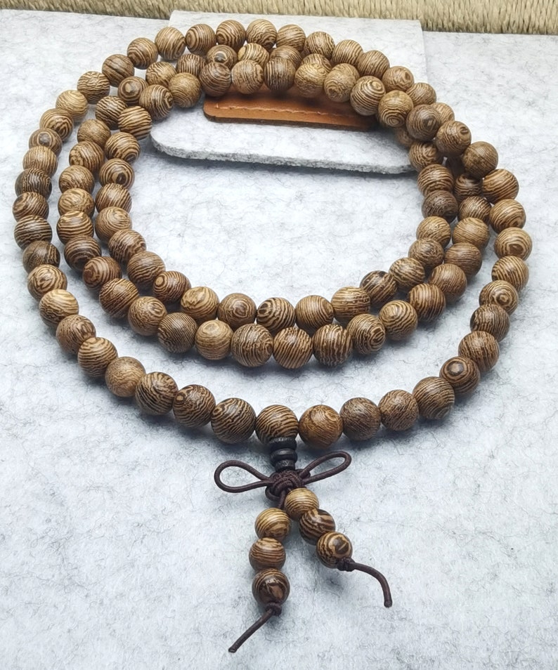 Puede incluir: Un collar de cuentas de oraci&oacute;n de madera con un acabado marr&oacute;n oscuro. Las cuentas son redondas y tienen un patr&oacute;n de vetas de madera visible. Una borla de cuentas m&aacute;s peque&ntilde;as y un cord&oacute;n anudado est&aacute;n unidos a la parte inferior del collar. Las cuentas se muestran sobre una superficie gris claro.