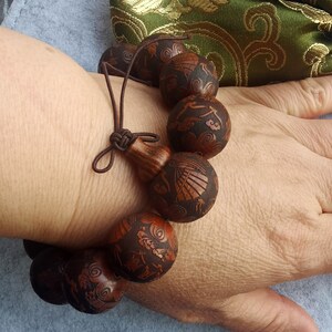 May include: A brown wooden bracelet with intricate carvings of clouds and birds. The bracelet is made of 10 beads and has a brown cord tie.