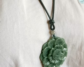 Puede incluir: Un collar con un colgante de jade verde en forma de flor en un cordón gris oscuro. El colgante está tallado en forma de flor con múltiples capas de pétalos.