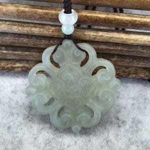 Peut inclure: Un pendentif en jade translucide vert p&acirc;le pr&eacute;sentant un motif complexe et sym&eacute;trique de volutes ajour&eacute;es. Le pendentif est suspendu &agrave; un cordon tress&eacute; marron fonc&eacute;, agr&eacute;ment&eacute; de deux petites perles rondes de couleur claire au-dessus.