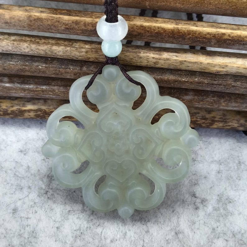 Peut inclure: Un pendentif en jade translucide vert p&acirc;le pr&eacute;sentant un motif complexe et sym&eacute;trique de volutes ajour&eacute;es. Le pendentif est suspendu &agrave; un cordon tress&eacute; marron fonc&eacute;, agr&eacute;ment&eacute; de deux petites perles rondes de couleur claire au-dessus.
