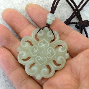 Peut inclure: Un pendentif en pierre sculpt&eacute;e translucide vert p&acirc;le pr&eacute;sentant un motif sym&eacute;trique ajour&eacute; de volutes et de c&oelig;urs. Le pendentif est suspendu &agrave; un cordon tress&eacute; marron fonc&eacute;, agr&eacute;ment&eacute; d'une petite perle vert clair au-dessus de la pi&egrave;ce principale.