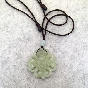 Peut inclure: Un pendentif en jade vert p&acirc;le pr&eacute;sentant un design complexe et sym&eacute;trique &agrave; volutes ajour&eacute;es. Le pendentif est suspendu &agrave; un collier en cordon marron fonc&eacute; r&eacute;glable, incluant une petite perle vert clair au-dessus de la pi&egrave;ce principale.