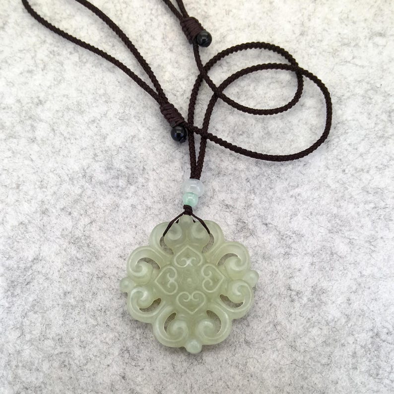 Peut inclure: Un pendentif en jade vert p&acirc;le pr&eacute;sentant un design complexe et sym&eacute;trique &agrave; volutes ajour&eacute;es. Le pendentif est suspendu &agrave; un collier en cordon marron fonc&eacute; r&eacute;glable, incluant une petite perle vert clair au-dessus de la pi&egrave;ce principale.