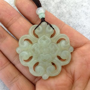 Peut inclure: Un pendentif en jade vert p&acirc;le pr&eacute;sentant un design ajour&eacute; complexe avec des motifs tourbillonnants sym&eacute;triques et une fleur centrale. Le pendentif est suspendu &agrave; un cordon marron fonc&eacute; orn&eacute; d'une petite perle vert clair translucide.