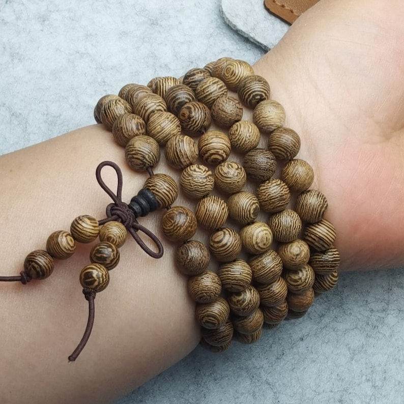 Puede incluir: Una pulsera de cuentas de madera con un patr&oacute;n de vetas de madera natural. La pulsera est&aacute; hecha de muchas cuentas redondas, ensartadas con un cord&oacute;n marr&oacute;n. Las cuentas son de color marr&oacute;n claro con vetas marrones m&aacute;s oscuras. La pulsera est&aacute; atada con un nudo decorativo.