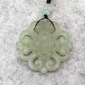 Peut inclure: Un pendentif en pierre translucide vert p&acirc;le pr&eacute;sentant un design ajour&eacute; sym&eacute;trique et complexe avec des motifs en forme de c&oelig;ur. Le pendentif est suspendu &agrave; un cordon marron fonc&eacute; orn&eacute; d'une petite perle vert clair au-dessus de la pi&egrave;ce.