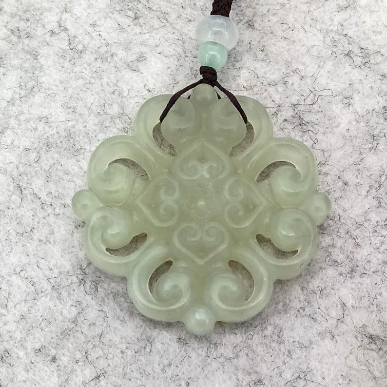 Peut inclure: Un pendentif en pierre translucide vert p&acirc;le pr&eacute;sentant un design ajour&eacute; sym&eacute;trique et complexe avec des motifs en forme de c&oelig;ur. Le pendentif est suspendu &agrave; un cordon marron fonc&eacute; orn&eacute; d'une petite perle vert clair au-dessus de la pi&egrave;ce.