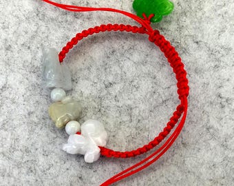 May include: A red cord bracelet featuring a series of carved stone charms in shades of white, gray, and tan, interspersed with small white beads. A bright green carved leaf-shaped charm is attached to the top, and the ends have small silver beads.