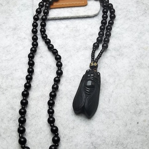Puede incluir: Un collar de cuentas negras con un colgante de jade negro tallado en forma de cigarra. El colgante tiene detalles dorados y caracteres chinos.
