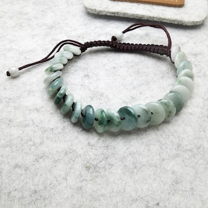 Könnte beinhalten: Ein Jade-Armband mit flachen, kreisförmigen Jadeperlen in Grün- und Weißtönen. Die Perlen sind auf einer verstellbaren, dunkelbraunen Kordel mit Schiebknotenverschluss und weißen Perlenakzenten aufgefädelt. Das Armband ist für das Handgelenk konzipiert.