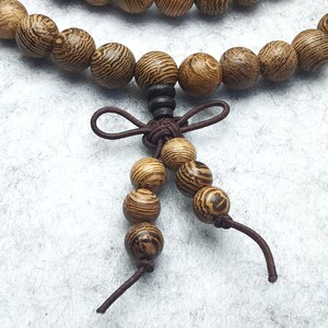 Puede incluir: Primer plano de una pulsera de cuentas de madera con una borla decorativa. Las cuentas son redondas, con un patr&oacute;n de vetas de madera natural en tonos marr&oacute;n y beige. La borla es de cord&oacute;n marr&oacute;n.