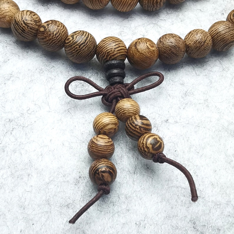 Puede incluir: Primer plano de una pulsera de cuentas de madera con una borla decorativa. Las cuentas son redondas, con un patr&oacute;n de vetas de madera natural en tonos marr&oacute;n y beige. La borla es de cord&oacute;n marr&oacute;n.