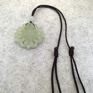 Peut inclure: Un pendentif en jade sculpt&eacute; vert p&acirc;le pr&eacute;sentant un motif sym&eacute;trique complexe en forme de n&oelig;ud. Le pendentif est suspendu &agrave; un cordon tress&eacute; marron fonc&eacute; comprenant deux petites perles coulissantes noires et une perle vert clair.