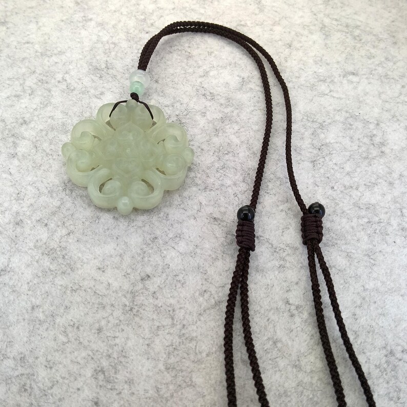 Peut inclure: Un pendentif en jade sculpt&eacute; vert p&acirc;le pr&eacute;sentant un motif sym&eacute;trique complexe en forme de n&oelig;ud. Le pendentif est suspendu &agrave; un cordon tress&eacute; marron fonc&eacute; comprenant deux petites perles coulissantes noires et une perle vert clair.