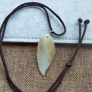 Puede incluir: Un colgante de jade blanco en forma de hoja en un collar de cordón marrón. El colgante está tallado en forma de hoja con vetas detalladas.