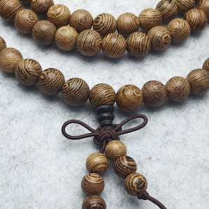 Puede incluir: Primer plano de un collar de cuentas de madera con un cord&oacute;n marr&oacute;n y un nudo decorativo. Las cuentas redondas son de color marr&oacute;n claro con un patr&oacute;n de vetas de madera a rayas. El collar se muestra sobre una superficie gris claro.