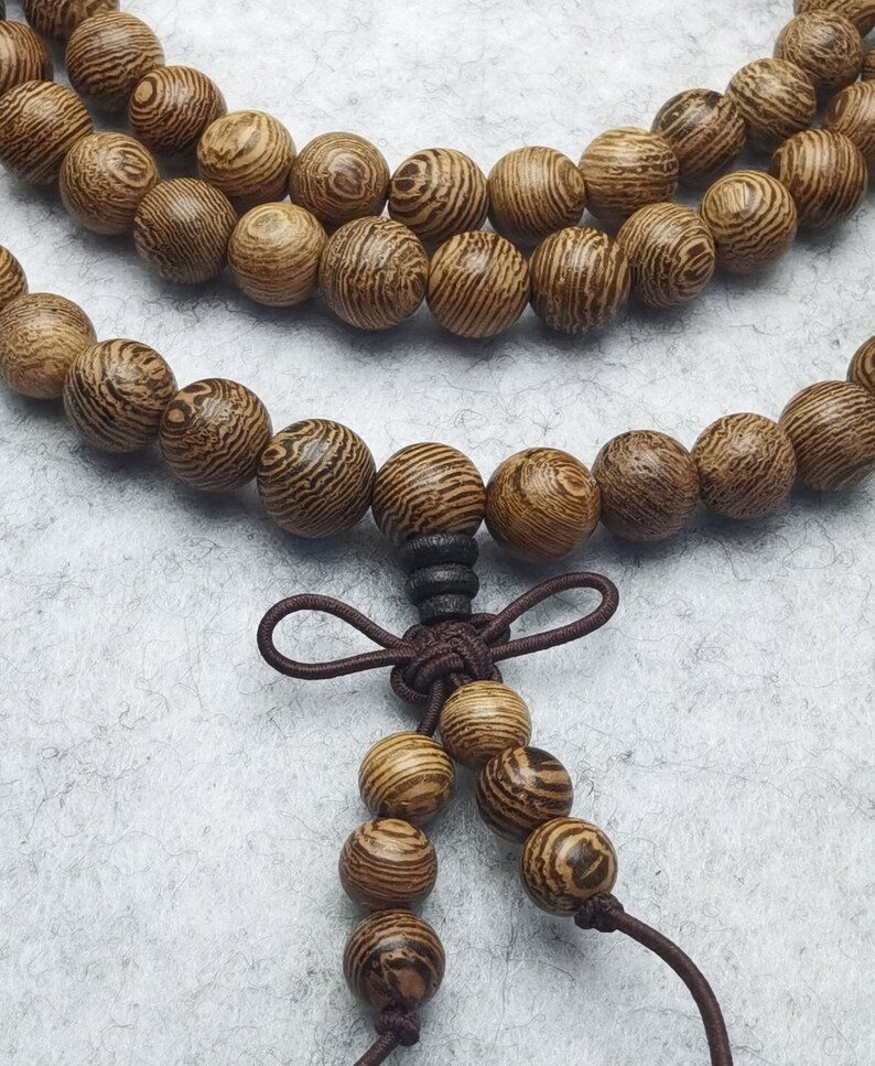 Puede incluir: Primer plano de un collar de cuentas de madera con un cord&oacute;n marr&oacute;n y un nudo decorativo. Las cuentas redondas son de color marr&oacute;n claro con un patr&oacute;n de vetas de madera a rayas. El collar se muestra sobre una superficie gris claro.