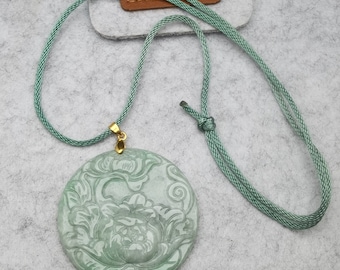 Puede incluir: Colgante redondo de jade verde pálido con un diseño floral tallado, suspendido de un cordón verde a juego. El colgante está sujeto a una fianza dorada. El cordón es ajustable. El fondo es gris claro.