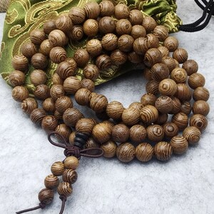 Puede incluir: Un collar de cuentas de oraci&oacute;n de madera con una borla marr&oacute;n y una bolsa verde con cord&oacute;n. Las cuentas son redondas y tienen un patr&oacute;n de vetas de madera. La bolsa es verde con un dise&ntilde;o floral dorado.