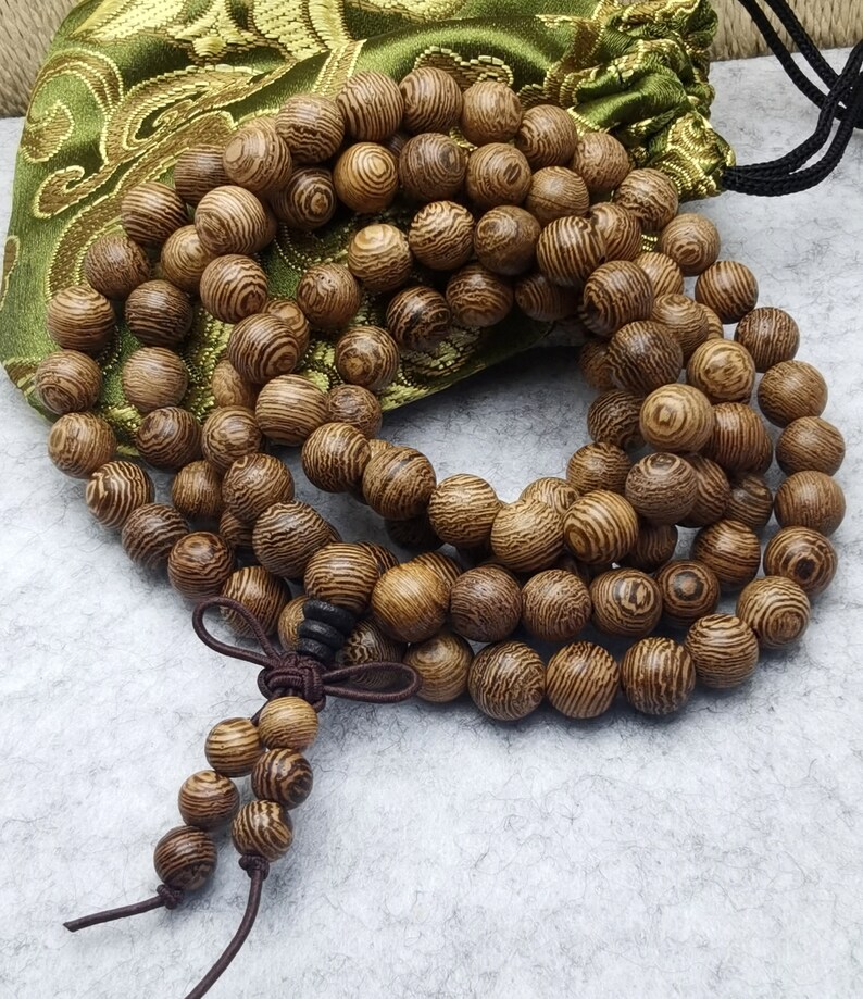 Puede incluir: Un collar de cuentas de oraci&oacute;n de madera con una borla marr&oacute;n y una bolsa verde con cord&oacute;n. Las cuentas son redondas y tienen un patr&oacute;n de vetas de madera. La bolsa es verde con un dise&ntilde;o floral dorado.