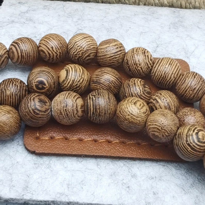 Puede incluir: Primer plano de cuentas de madera con un patr&oacute;n a rayas en tonos marrones y beige. Las cuentas redondas est&aacute;n dispuestas sobre una superficie de cuero marr&oacute;n, lo que sugiere un tema artesanal o de joyer&iacute;a.