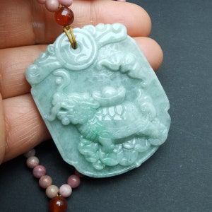 May include: A jade pendant necklace featuring a green jade carving of a dragon and a turtle. The pendant is intricately detailed and has a gold-colored cord.