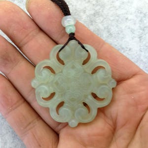 Peut inclure: Un pendentif en jade vert p&acirc;le pr&eacute;sentant un motif ajour&eacute; complexe et sym&eacute;trique en forme de volutes. Le pendentif est suspendu &agrave; un cordon marron fonc&eacute;, agr&eacute;ment&eacute; d'une petite perle translucide vert clair plac&eacute;e au-dessus de la pi&egrave;ce.