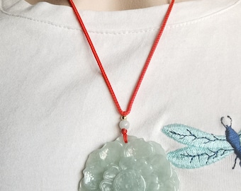May include: A pale green, translucent, circular pendant carved into a multi-layered floral shape. The pendant hangs from a thin, bright red cord, accented by a small, round, white bead and a tiny gold-toned bead just above the floral carving.
