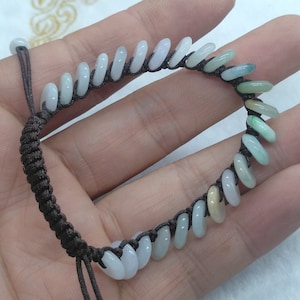 Puede incluir: Un brazalete de jade con un cordón marrón. El brazalete está hecho de pequeñas cuentas redondas de jade que están enhebradas en un cordón marrón. Las cuentas son de varios tonos de verde, blanco y amarillo.