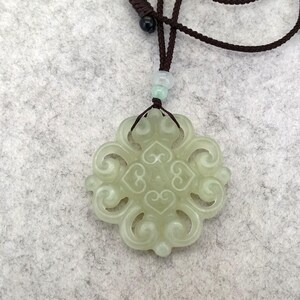 Peut inclure: Un pendentif en jade vert p&acirc;le pr&eacute;sentant un design ajour&eacute; complexe compos&eacute; de volutes sym&eacute;triques et de motifs en forme de c&oelig;ur. Le pendentif est suspendu &agrave; un cordon marron fonc&eacute;, incluant une petite perle claire au-dessus de la pierre.
