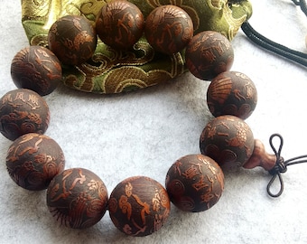 Puede incluir: Un brazalete de cuentas de madera marrón con tallas intrincadas. Las cuentas son redondas y tienen un acabado brillante. El brazalete está atado con un cordón y una borla marrones.