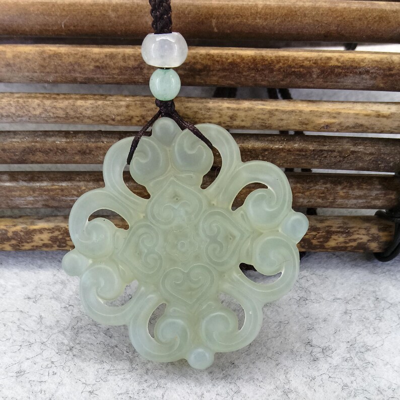 Peut inclure: Un pendentif en jade translucide vert p&acirc;le pr&eacute;sentant un design ajour&eacute; sym&eacute;trique et complexe avec des motifs en forme de c&oelig;ur tourbillonnants. Le pendentif est suspendu &agrave; un cordon marron fonc&eacute; orn&eacute; de deux petites perles rondes claires.