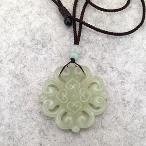 Peut inclure: Un pendentif en pierre translucide vert p&acirc;le pr&eacute;sentant un motif ajour&eacute; complexe et sym&eacute;trique en forme de volutes. Le pendentif est suspendu &agrave; un cordon marron fonc&eacute; avec un n&oelig;ud coulissant r&eacute;glable et deux petites perles claires au-dessus.