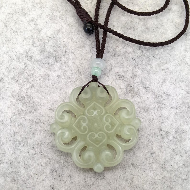 Peut inclure: Un pendentif en pierre translucide vert p&acirc;le pr&eacute;sentant un motif ajour&eacute; complexe et sym&eacute;trique en forme de volutes. Le pendentif est suspendu &agrave; un cordon marron fonc&eacute; avec un n&oelig;ud coulissant r&eacute;glable et deux petites perles claires au-dessus.