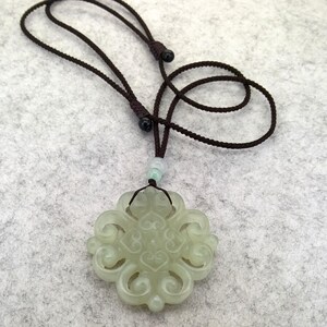 Peut inclure: Un pendentif en jade vert p&acirc;le pr&eacute;sentant un design complexe &agrave; volutes ajour&eacute;es, suspendu &agrave; un cordon r&eacute;glable marron fonc&eacute;. Le pendentif est agr&eacute;ment&eacute; d'une petite perle vert clair translucide plac&eacute;e juste au-dessus de la pierre sculpt&eacute;e.