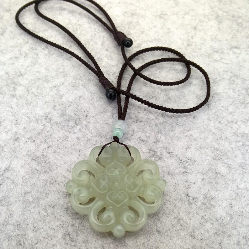Peut inclure: Un pendentif en jade vert p&acirc;le pr&eacute;sentant un design complexe &agrave; volutes ajour&eacute;es, suspendu &agrave; un cordon r&eacute;glable marron fonc&eacute;. Le pendentif est agr&eacute;ment&eacute; d'une petite perle vert clair translucide plac&eacute;e juste au-dessus de la pierre sculpt&eacute;e.
