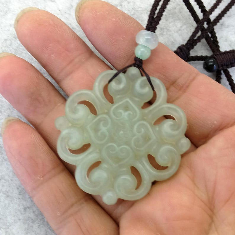 Peut inclure: Un pendentif en pierre translucide vert p&acirc;le pr&eacute;sentant un design ajour&eacute; sym&eacute;trique et complexe avec des motifs en forme de c&oelig;ur tourbillonnants. Le pendentif est suspendu &agrave; un cordon marron fonc&eacute; avec une petite perle claire.