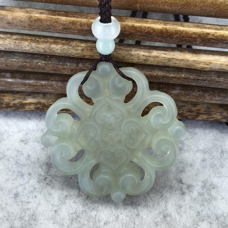 Peut inclure: Un pendentif en jade translucide vert p&acirc;le pr&eacute;sentant un design ajour&eacute; sym&eacute;trique et complexe avec des motifs en forme de c&oelig;ur tourbillonnants. Le pendentif est suspendu &agrave; un cordon tress&eacute; marron fonc&eacute;, orn&eacute; d'une petite perle ronde claire.