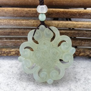 Peut inclure: Un pendentif en pierre translucide vert p&acirc;le pr&eacute;sentant un design complexe &agrave; volutes ajour&eacute;es. Le pendentif est suspendu &agrave; un cordon marron fonc&eacute; orn&eacute; de deux petites perles rondes de couleur claire situ&eacute;es au-dessus de la pi&egrave;ce sculpt&eacute;e.