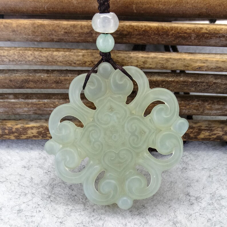 Peut inclure: Un pendentif en pierre translucide vert p&acirc;le pr&eacute;sentant un design complexe &agrave; volutes ajour&eacute;es. Le pendentif est suspendu &agrave; un cordon marron fonc&eacute; orn&eacute; de deux petites perles rondes de couleur claire situ&eacute;es au-dessus de la pi&egrave;ce sculpt&eacute;e.