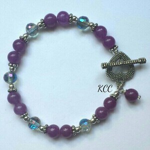 May include: Purple and silver beaded bracelet with a heart-shaped clasp. The bracelet features alternating purple beads and iridescent clear beads.